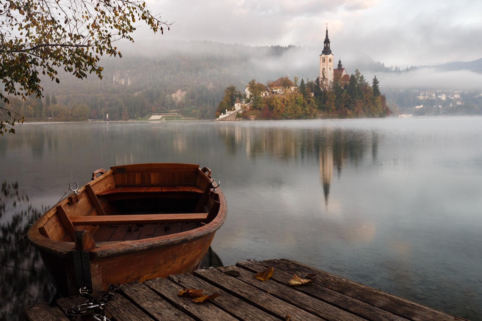 Image of a wooden dingy boat, moored to a wooden deck. Across a body of water is a mystical building, including a steeple, surrounded by trees. There is reflection of the trees and building in the water.
